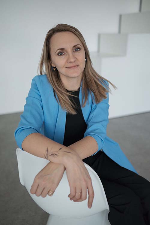 Irina Wengenroth aus Bad Nauheim schaut in die Kamera, sie hat einen blauen Blazer an und sitzt freundlich auf einem weißen Stuhl